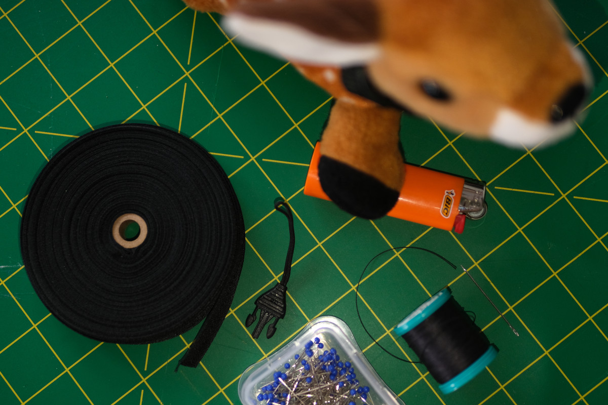 An assortment of crafting materials and tools - a small buckle, pins and a needle, thread, a roll of nylon webbing, and small deer plush stealing a lighter